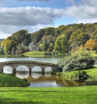 A picturesque landscape featuring a calm lake surrounded by lush, green hills and trees with hints of autumn foliage. A stone arch bridge crosses a narrow part of the water in the foreground, and a small, classical-style temple with a dome is visible on the far bank. The sky is partly cloudy.