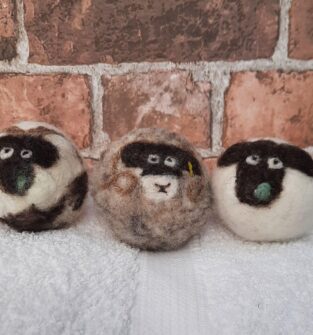 A close-up of three handmade, spherical wool crafts, designed to resemble sheep faces. The wool 'sheep' have dark faces, white eyes, and small green felt noses. They are sitting side-by-side on a white, terry-cloth towel in front of a rustic red brick wall.