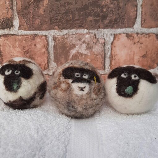 A close-up of three handmade, spherical wool crafts, designed to resemble sheep faces. The wool 'sheep' have dark faces, white eyes, and small green felt noses. They are sitting side-by-side on a white, terry-cloth towel in front of a rustic red brick wall.