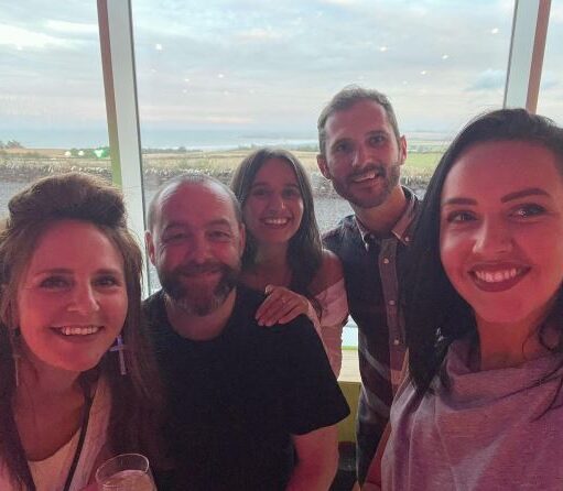 A smiling group of five adults, three women and two men, posing for a selfie indoors in front of a large window. The background visible through the window is an expansive, slightly overcast coastal landscape at what appears to be either dawn or dusk, with the sky a soft pink and grey. The people in the group are closely gathered and appear happy, with the lighting casting a warm, slightly reddish glow over their faces. The woman on the far right holds the camera for the selfie.