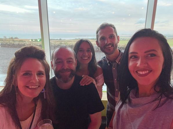 A smiling group of five adults, three women and two men, posing for a selfie indoors in front of a large window. The background visible through the window is an expansive, slightly overcast coastal landscape at what appears to be either dawn or dusk, with the sky a soft pink and grey. The people in the group are closely gathered and appear happy, with the lighting casting a warm, slightly reddish glow over their faces. The woman on the far right holds the camera for the selfie.