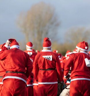 A large crowd of people, viewed from behind, are all wearing full Santa suits—red jackets, trousers, and hats with white trim—suggesting a Santa Fun Run or Charity event. The photograph is taken outdoors under a slightly overcast sky, with the focus on the backs of the participants who are gathered closely together. One participant in the center has a race number "60" pinned to the back of their suit, and the person on the right has a white race bib hanging down. The background is blurred, showing bare trees against the pale sky and hints of a path or field in front of them.