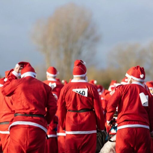 A large crowd of people, viewed from behind, are all wearing full Santa suits—red jackets, trousers, and hats with white trim—suggesting a Santa Fun Run or Charity event. The photograph is taken outdoors under a slightly overcast sky, with the focus on the backs of the participants who are gathered closely together. One participant in the center has a race number "60" pinned to the back of their suit, and the person on the right has a white race bib hanging down. The background is blurred, showing bare trees against the pale sky and hints of a path or field in front of them.