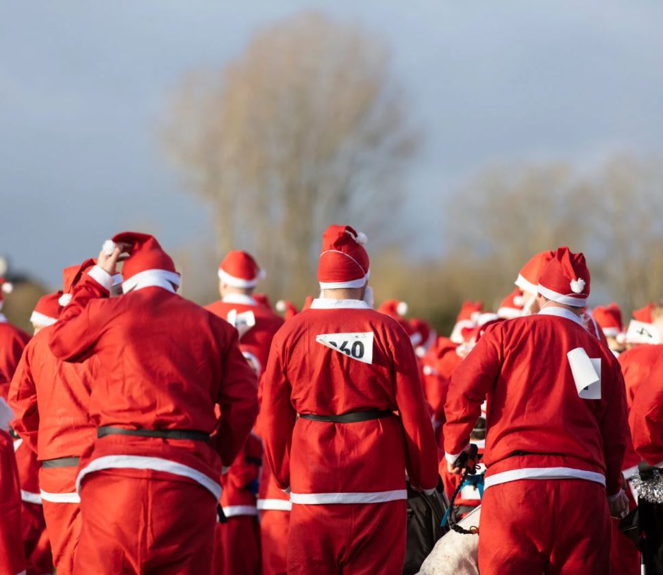 A large crowd of people, viewed from behind, are all wearing full Santa suits—red jackets, trousers, and hats with white trim—suggesting a Santa Fun Run or Charity event. The photograph is taken outdoors under a slightly overcast sky, with the focus on the backs of the participants who are gathered closely together. One participant in the center has a race number "60" pinned to the back of their suit, and the person on the right has a white race bib hanging down. The background is blurred, showing bare trees against the pale sky and hints of a path or field in front of them.