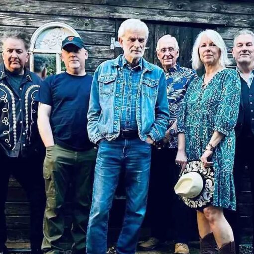 A group of six musicians, consisting of one woman and five men, stand outdoors posing for a band photo against a weathered, rustic wooden shed backdrop. All members are adults, wearing a variety of casual and slightly Western-inspired attire. The man in the center wears a full denim outfit—jeans and a jacket—and has white hair. The woman on the right wears a patterned teal-green dress and brown boots, holding a white Western hat. The overall lighting is dim and slightly cool, giving the image a moody, rustic feel.