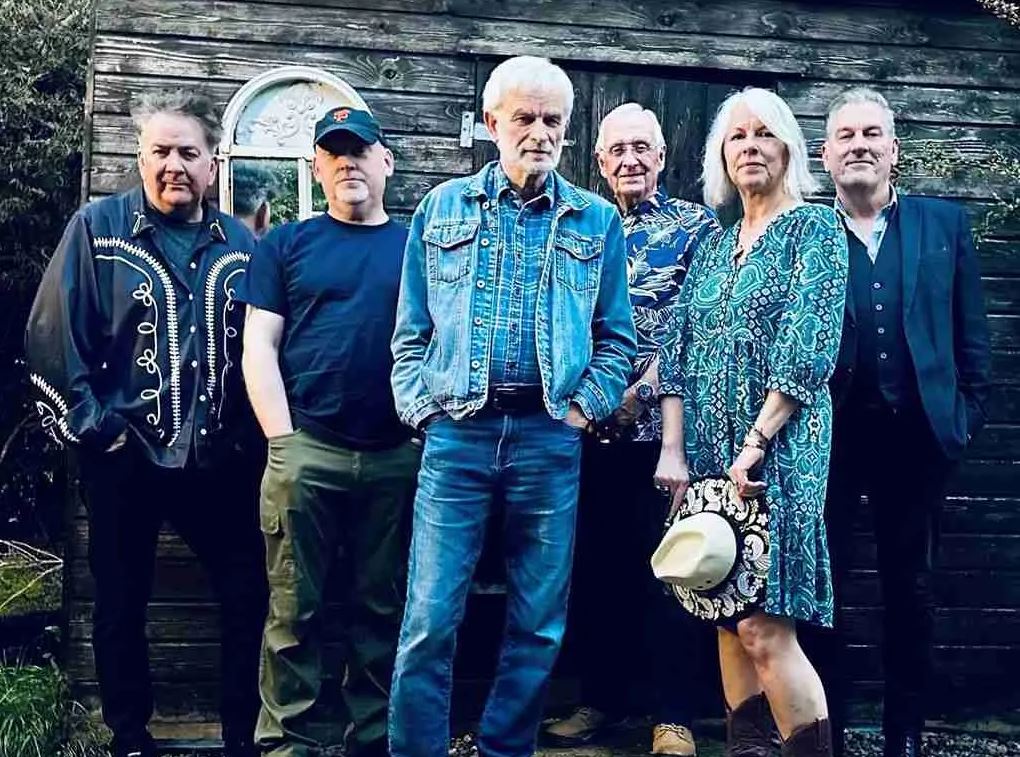 A group of six musicians, consisting of one woman and five men, stand outdoors posing for a band photo against a weathered, rustic wooden shed backdrop. All members are adults, wearing a variety of casual and slightly Western-inspired attire. The man in the center wears a full denim outfit—jeans and a jacket—and has white hair. The woman on the right wears a patterned teal-green dress and brown boots, holding a white Western hat. The overall lighting is dim and slightly cool, giving the image a moody, rustic feel.