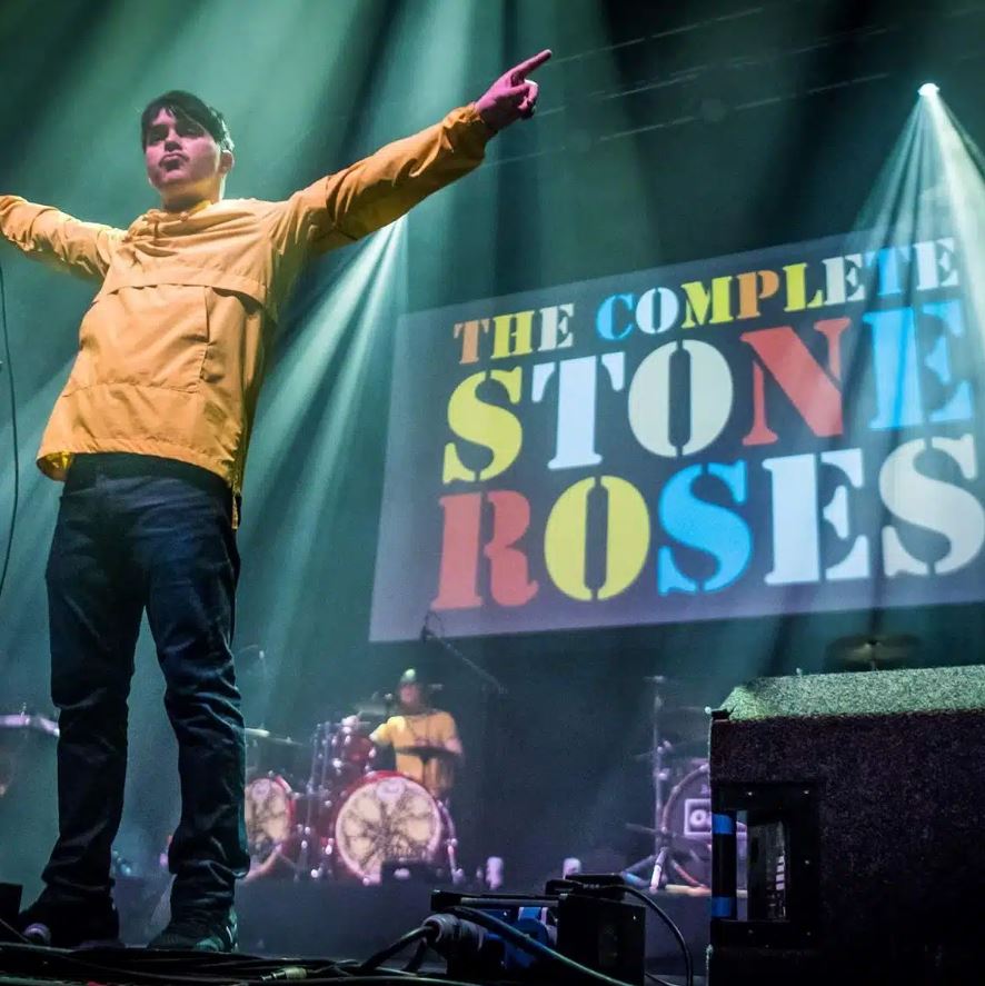 A live music performance on a brightly lit stage, with the lead singer of a tribute band standing prominently in the foreground. The performer, a young man, wears a mustard-yellow cagoule-style jacket and dark trousers, with his arms outstretched and fingers pointing up. Behind him, a large screen displays the name of the act: "THE COMPLETE STONE ROSES" with the word "STONE" in yellow and "ROSES" in red block letters. Stage lights create dramatic beams of white light, and in the background, a drummer is visible seated behind a drum kit with a distinctive logo on the bass drum head.