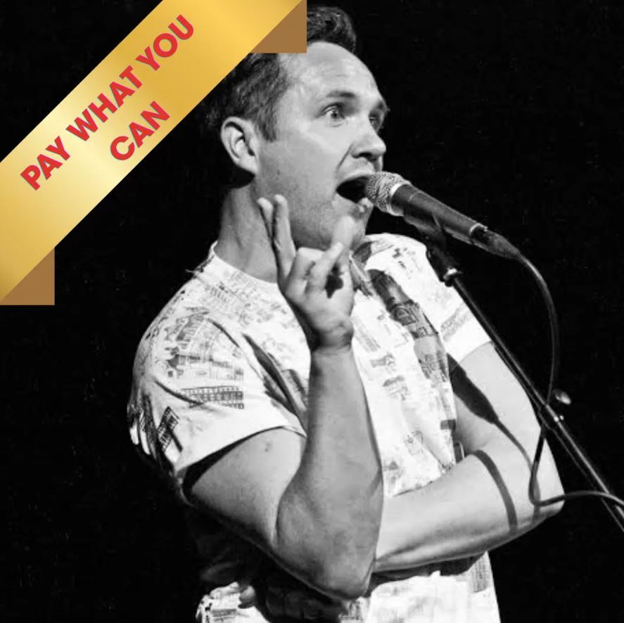 A black and white photograph of a male comedian or performer on a dark stage, captured mid-performance while speaking into a microphone. The man is wearing a patterned or graphic t-shirt, gesturing with one hand held up in a pose resembling a rock-and-roll or shock sign, and looking up and to the right with an open mouth. A decorative gold and yellow banner overlaid in the top left corner features the text "PAY WHAT YOU CAN" in red capital letters.