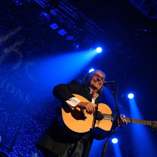 A live concert photo features an adult male musician, possibly a folk or rock artist, smiling broadly as he plays an acoustic guitar on a stage bathed in dramatic blue spotlighting. The performer is wearing glasses, a white collared shirt, and a dark blazer, with his long hair flowing back, illuminated by the bright beams of light shining down from above. The background is dark, with specks of light creating a sense of a packed venue or digital effect, and the neck of another guitar is visible in the lower left corner.