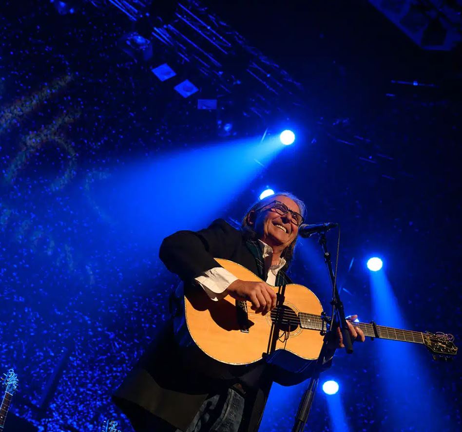 A live concert photo features an adult male musician, possibly a folk or rock artist, smiling broadly as he plays an acoustic guitar on a stage bathed in dramatic blue spotlighting. The performer is wearing glasses, a white collared shirt, and a dark blazer, with his long hair flowing back, illuminated by the bright beams of light shining down from above. The background is dark, with specks of light creating a sense of a packed venue or digital effect, and the neck of another guitar is visible in the lower left corner.
