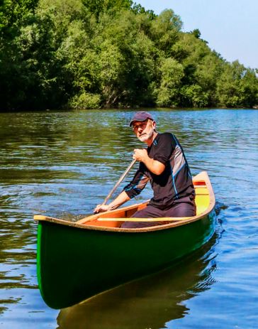A middle-aged or older man wearing a dark shirt and a cap is paddling a bright green canoe on a calm body of water. He is seated and actively paddling on the right side of the canoe, looking directly at the camera. The background consists of a dense, lush green forest meeting the water under a bright sky.
