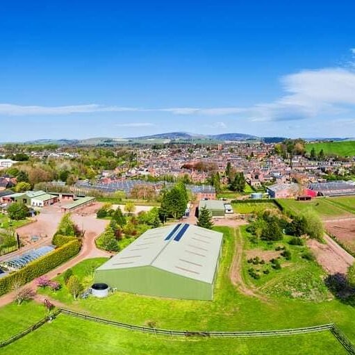 An aerial view shows a large, light green agricultural building (likely a barn or shed) in the foreground, with solar panels on its roof, surrounded by fenced grassy fields and other smaller farm structures. In the background, there is a town with dense housing, leading up to rolling green hills under a bright blue sky with some white clouds.