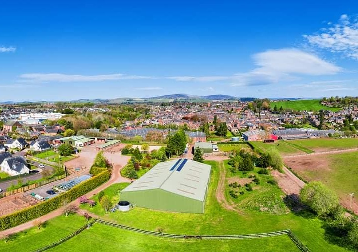 An aerial view shows a large, light green agricultural building (likely a barn or shed) in the foreground, with solar panels on its roof, surrounded by fenced grassy fields and other smaller farm structures. In the background, there is a town with dense housing, leading up to rolling green hills under a bright blue sky with some white clouds.