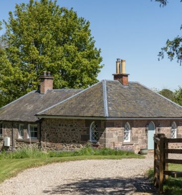 Exterior view of a charming stone cottage with a dark roof and a gravel driveway in bright sunlight.