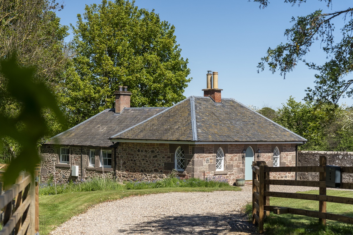Exterior view of a charming stone cottage with a dark roof and a gravel driveway in bright sunlight.