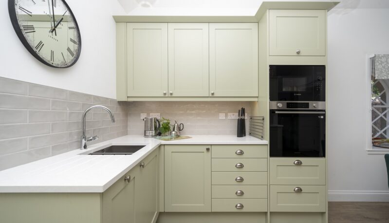 Contemporary kitchen with sage green cabinets, white countertops, a large wall clock, and a stainless steel sink.