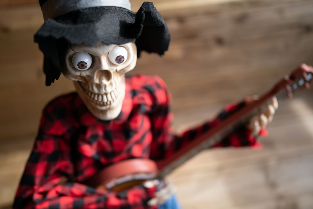 A spooky skeleton decoration wearing a black top hat and a red and black plaid shirt is depicted playing a small guitar. The background is wooden panelling.