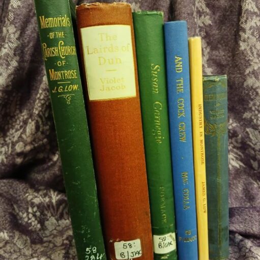 A collection of seven vintage books standing upright against a dark purple, patterned fabric background. The visible titles include "Memorials of the Parish Church of Montrose" (dark green), "The Lairds of Dun" by Violet Jacob (tall, reddish-brown spine), "Susan Carnegie" (green), "And The Cook Crew" (blue), and "Industry in Montrose" (yellow). Some of the spines have library or owner's labels near the bottom.