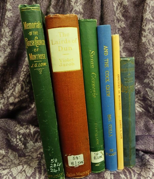 A collection of seven vintage books standing upright against a dark purple, patterned fabric background. The visible titles include "Memorials of the Parish Church of Montrose" (dark green), "The Lairds of Dun" by Violet Jacob (tall, reddish-brown spine), "Susan Carnegie" (green), "And The Cook Crew" (blue), and "Industry in Montrose" (yellow). Some of the spines have library or owner's labels near the bottom.