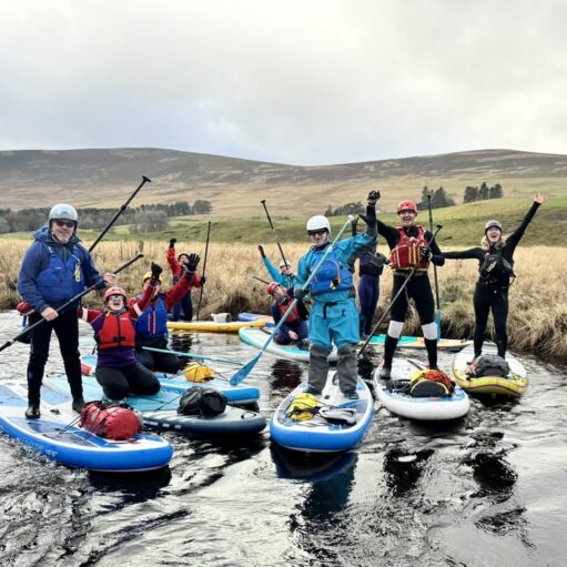 A group of eight people are standing and kneeling on inflatable paddleboards (SUPs) in a calm, shallow river. They are all wearing outdoor gear including helmets, wetsuits, and buoyancy aids, and most are raising their paddles or hands in a cheering gesture. The background shows a hilly, rural landscape with brown grasses and a cloudy sky.