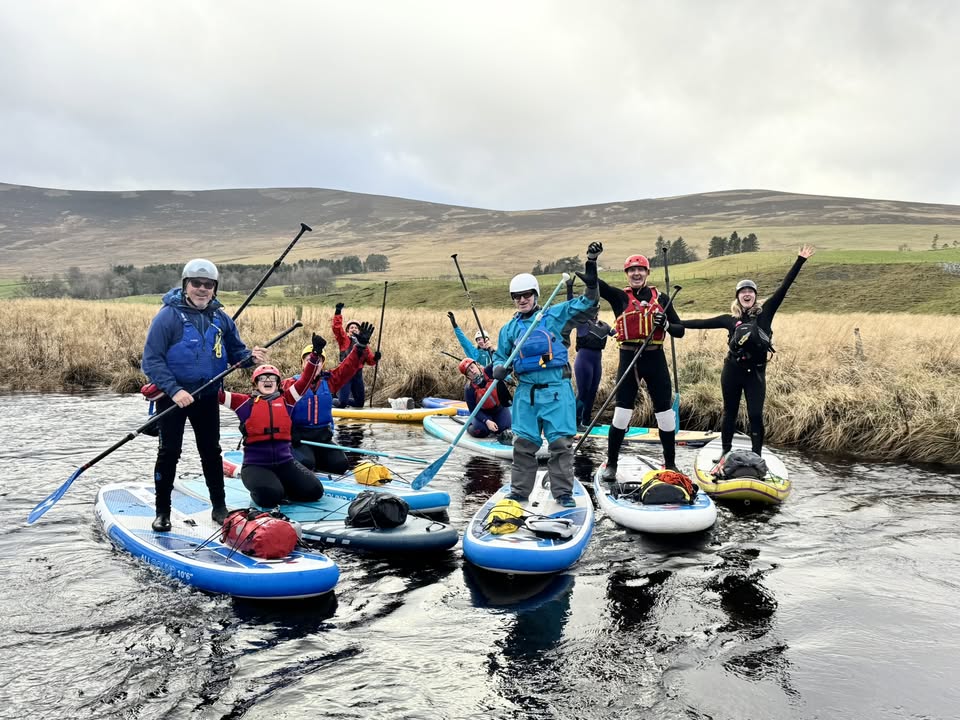 A group of eight people are standing and kneeling on inflatable paddleboards (SUPs) in a calm, shallow river. They are all wearing outdoor gear including helmets, wetsuits, and buoyancy aids, and most are raising their paddles or hands in a cheering gesture. The background shows a hilly, rural landscape with brown grasses and a cloudy sky.