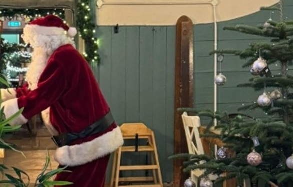 A photo showing a person dressed as Santa Claus wearing a red suit, hat, and a black belt, leaning over toward the left side of the frame. In the foreground on the right is a decorated Christmas tree with silver baubles. The background features a wooden panel wall painted a pale green or teal, with string lights and a wooden ladder.