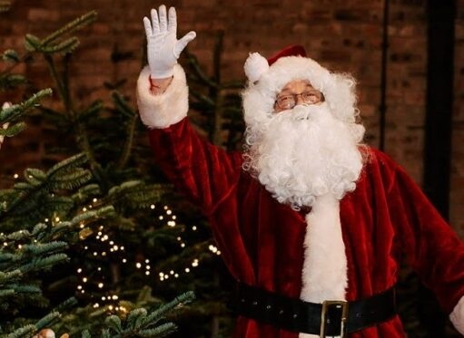 A photograph of a person dressed as Santa Claus in a red suit, white gloves, and hat, waving his right hand while standing next to a decorated Christmas tree. The background is a dark, rustic brick wall.