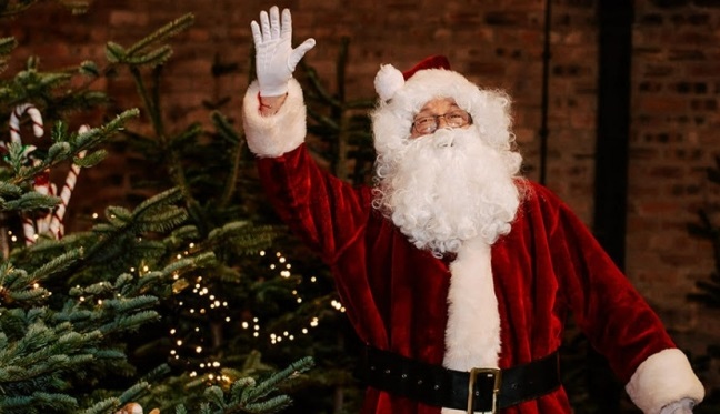 A photograph of a person dressed as Santa Claus in a red suit, white gloves, and hat, waving his right hand while standing next to a decorated Christmas tree. The background is a dark, rustic brick wall.