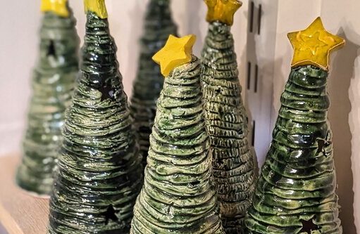 A close-up of several handmade ceramic Christmas trees. The trees are conical, textured, glazed in dark green, and each is topped with a distinct yellow star. They are displayed on a wooden shelf.