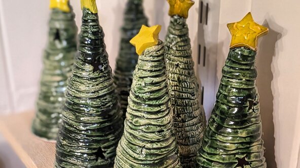 A close-up of several handmade ceramic Christmas trees. The trees are conical, textured, glazed in dark green, and each is topped with a distinct yellow star. They are displayed on a wooden shelf.