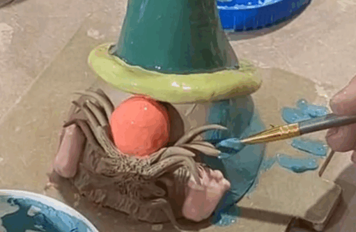 A close-up image showing a person's hand painting a ceramic gnome figure with a small paintbrush. The gnome has a large orange nose, brown clay beard details, a green conical hat with a yellow rim, and the person is applying a light blue glaze to the figure's body.
