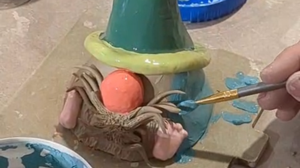 A close-up image showing a person's hand painting a ceramic gnome figure with a small paintbrush. The gnome has a large orange nose, brown clay beard details, a green conical hat with a yellow rim, and the person is applying a light blue glaze to the figure's body.