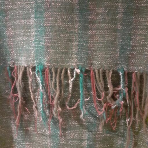 A close-up of a woven textile, likely a shawl or scarf, featuring a rough weave with vertical stripes in shades of moss green and teal. The bottom edge shows a frayed fringe with loose threads of green, teal, off-white, and light pink.