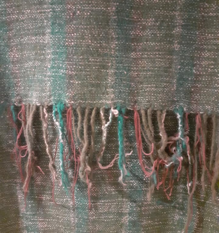 A close-up of a woven textile, likely a shawl or scarf, featuring a rough weave with vertical stripes in shades of moss green and teal. The bottom edge shows a frayed fringe with loose threads of green, teal, off-white, and light pink.