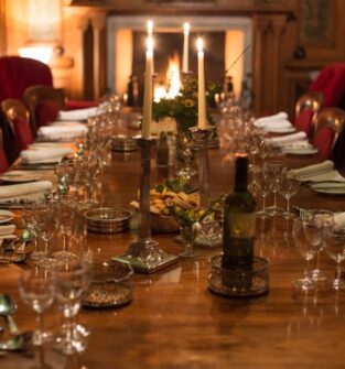 A dimly lit, formal dining room in a grand, wood-paneled setting. A long, polished wooden table is set for a dinner party with multiple place settings, wine glasses, white napkins, and silverware. The table is decorated with tall silver candelabras, a bottle of wine, and small bowls of nuts. Two red velvet wingback chairs flank a fireplace in the background, where a soft glow is visible.