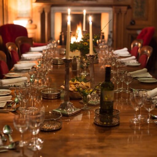 A dimly lit, formal dining room in a grand, wood-paneled setting. A long, polished wooden table is set for a dinner party with multiple place settings, wine glasses, white napkins, and silverware. The table is decorated with tall silver candelabras, a bottle of wine, and small bowls of nuts. Two red velvet wingback chairs flank a fireplace in the background, where a soft glow is visible.