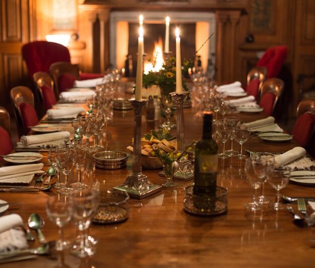 A dimly lit, formal dining room in a grand, wood-paneled setting. A long, polished wooden table is set for a dinner party with multiple place settings, wine glasses, white napkins, and silverware. The table is decorated with tall silver candelabras, a bottle of wine, and small bowls of nuts. Two red velvet wingback chairs flank a fireplace in the background, where a soft glow is visible.
