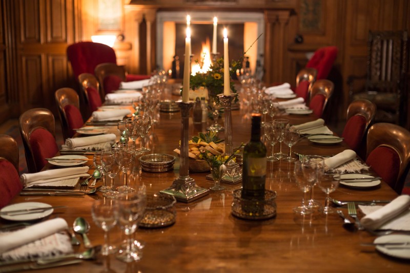 A dimly lit, formal dining room in a grand, wood-paneled setting. A long, polished wooden table is set for a dinner party with multiple place settings, wine glasses, white napkins, and silverware. The table is decorated with tall silver candelabras, a bottle of wine, and small bowls of nuts. Two red velvet wingback chairs flank a fireplace in the background, where a soft glow is visible.