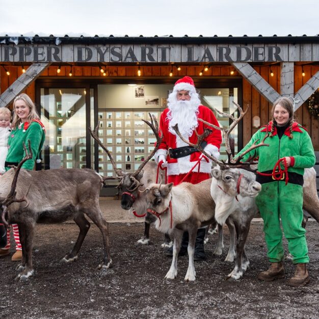 A holiday scene in front of the "Upper Dysart Larder" farm shop. Santa Claus stands with four reindeer being held on leashes. Two women in matching green elf outfits are also in the picture, one of whom is holding a baby dressed in white. The building behind them is made of wood with large glass doors and is decorated with string lights.