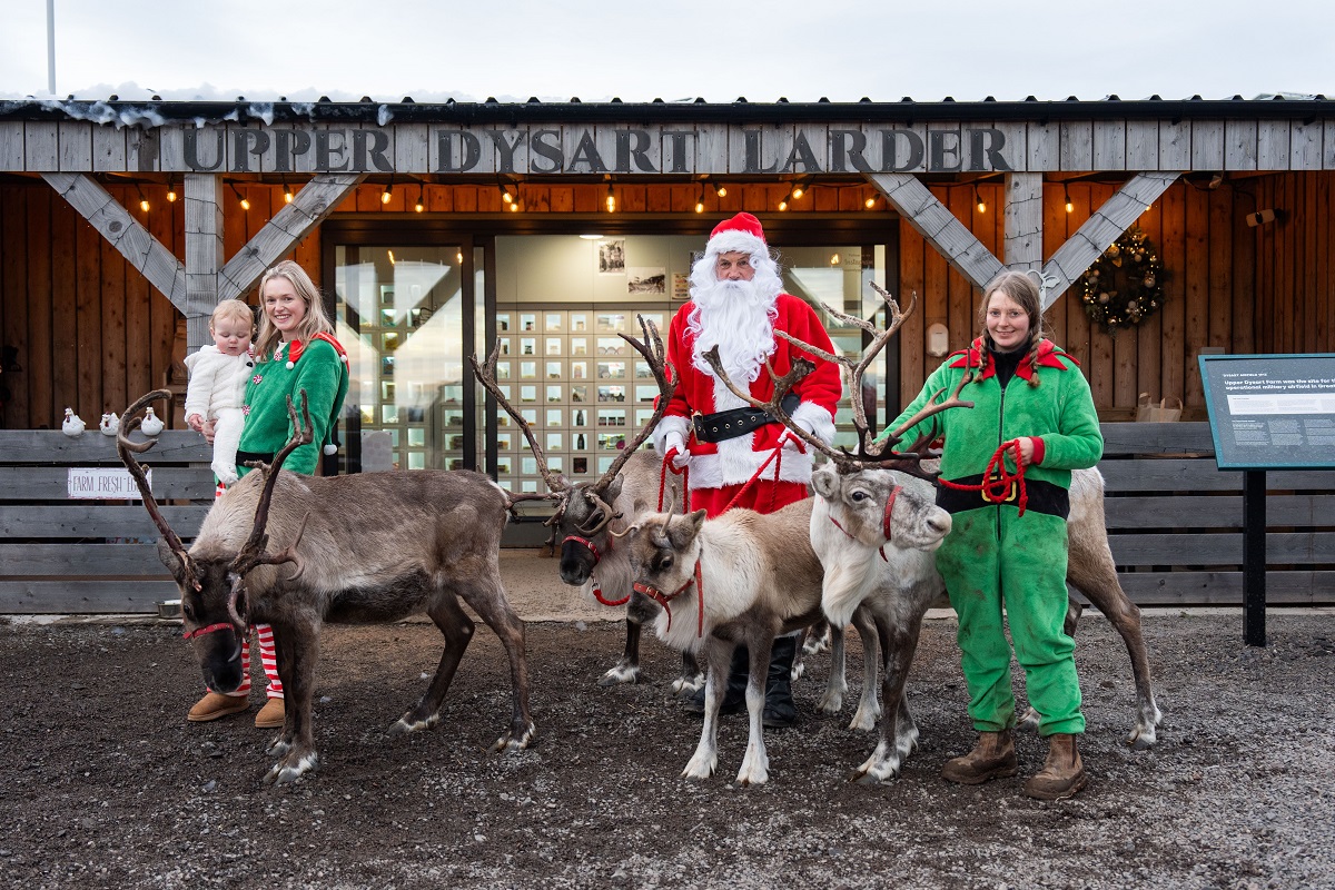 A holiday scene in front of the "Upper Dysart Larder" farm shop. Santa Claus stands with four reindeer being held on leashes. Two women in matching green elf outfits are also in the picture, one of whom is holding a baby dressed in white. The building behind them is made of wood with large glass doors and is decorated with string lights.