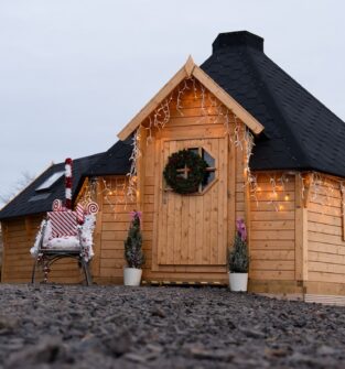 A cosy, small wooden cabin, likely a Santa's Grotto, with a dark, conical roof and decorated with a Christmas wreath on the door and twinkling lights along the eaves. A red and white sign on the left says "SANTA'S GROTTO". A decorative chair wrapped in candy-cane striped material sits outside the entrance, flanked by two small potted fir trees. The photo is taken from a low angle across a rocky ground.