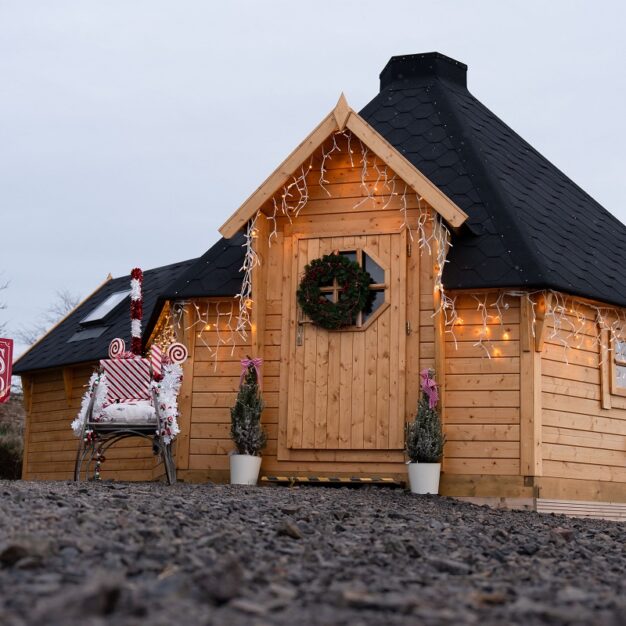 A cosy, small wooden cabin, likely a Santa's Grotto, with a dark, conical roof and decorated with a Christmas wreath on the door and twinkling lights along the eaves. A red and white sign on the left says "SANTA'S GROTTO". A decorative chair wrapped in candy-cane striped material sits outside the entrance, flanked by two small potted fir trees. The photo is taken from a low angle across a rocky ground.