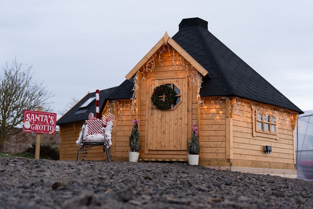 A cosy, small wooden cabin, likely a Santa's Grotto, with a dark, conical roof and decorated with a Christmas wreath on the door and twinkling lights along the eaves. A red and white sign on the left says "SANTA'S GROTTO". A decorative chair wrapped in candy-cane striped material sits outside the entrance, flanked by two small potted fir trees. The photo is taken from a low angle across a rocky ground.