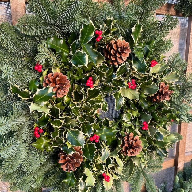 A close-up of a festive Christmas wreath made of variegated holly leaves and decorated with several pine cones and bright red berries. The holly wreath is mounted on a larger ring of dark green fir branches and set against a wooden background.