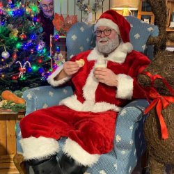 Santa Claus sitting in a patterned blue armchair, wearing his traditional red suit, holding a glass of milk and a cookie. A decorated Christmas tree is visible on the left, and a straw reindeer statue is on the right.
