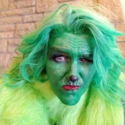 A close-up portrait of a person in elaborate, professional-quality Grinch makeup and costume. The person's face is painted green with wrinkles and shading, they have a red lipstick and a furrowed brow, and they are wearing a bright green wig and a lime-green fluffy coat. One eye appears to be a light color, possibly a contact lens.