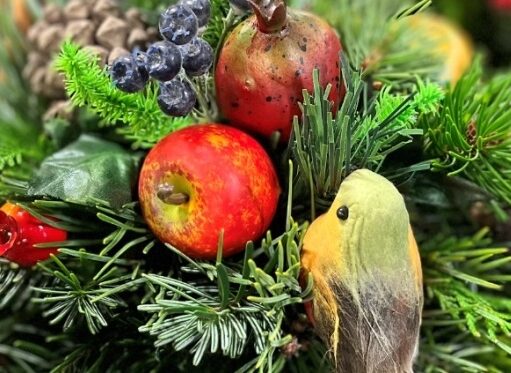 A close-up of a festive Christmas floral arrangement featuring a mix of evergreen branches, a red and yellow artificial apple, a small artificial pomegranate, dark blue berries, and a small decorative robin bird with a yellow breast.