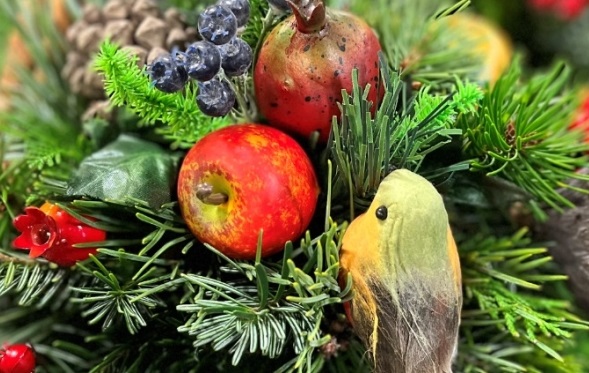 A close-up of a festive Christmas floral arrangement featuring a mix of evergreen branches, a red and yellow artificial apple, a small artificial pomegranate, dark blue berries, and a small decorative robin bird with a yellow breast.