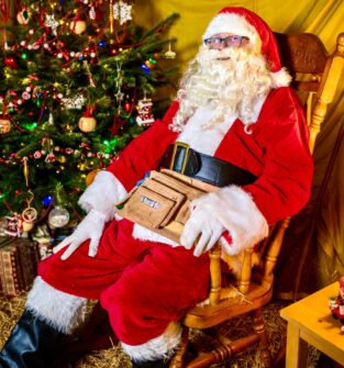 Santa Claus dressed in a traditional red suit with white fur trim, sitting in a wooden rocking chair beside a decorated Christmas tree. Santa has a large black belt and a tan utility pouch around the waist, with wrapped presents visible under the tree and festive decorations in the background.