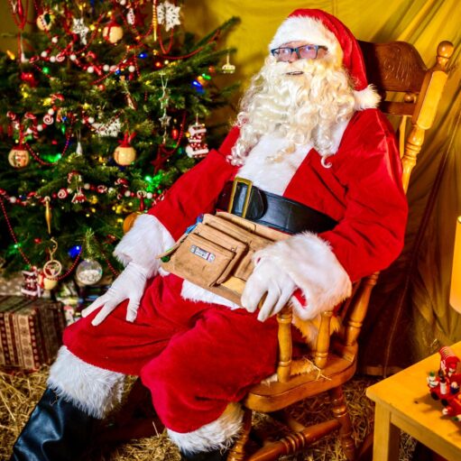 Santa Claus dressed in a traditional red suit with white fur trim, sitting in a wooden rocking chair beside a decorated Christmas tree. Santa has a large black belt and a tan utility pouch around the waist, with wrapped presents visible under the tree and festive decorations in the background.